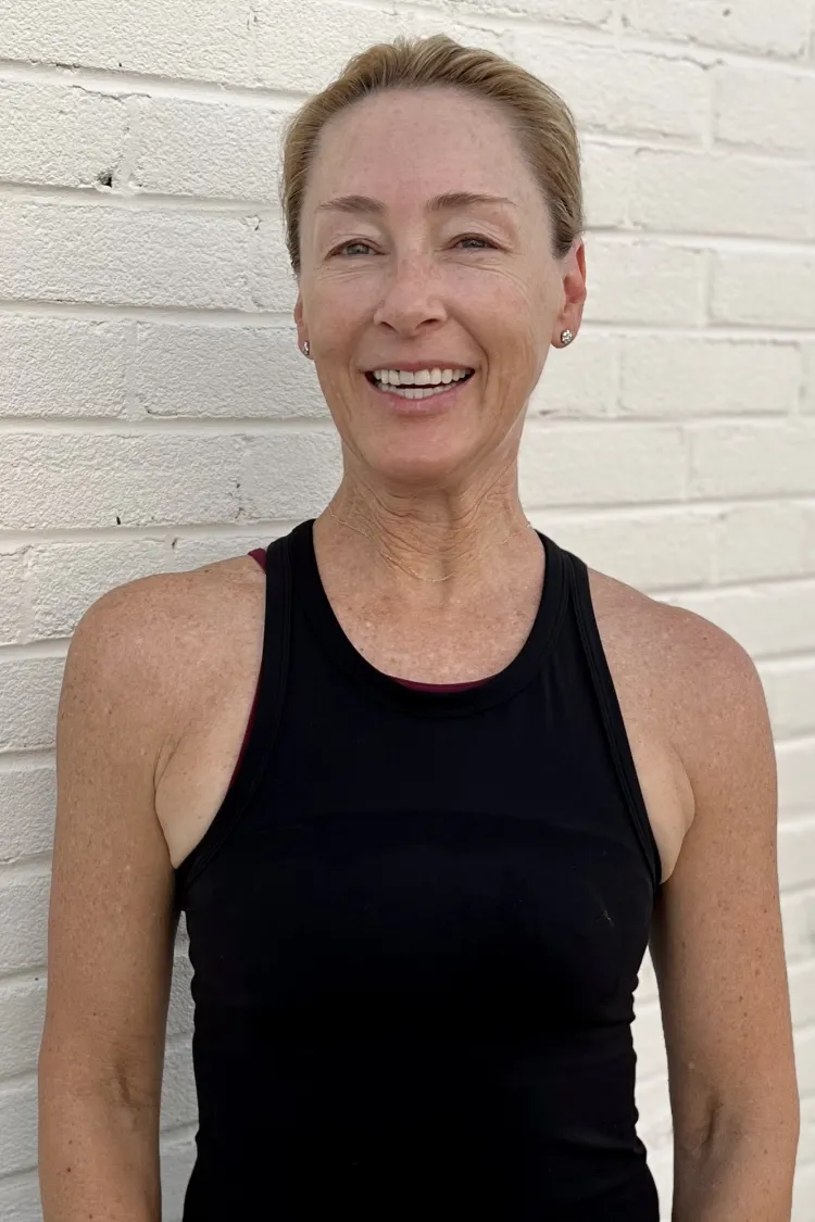 Smiling woman in a black tank top standing against a white brick wall.