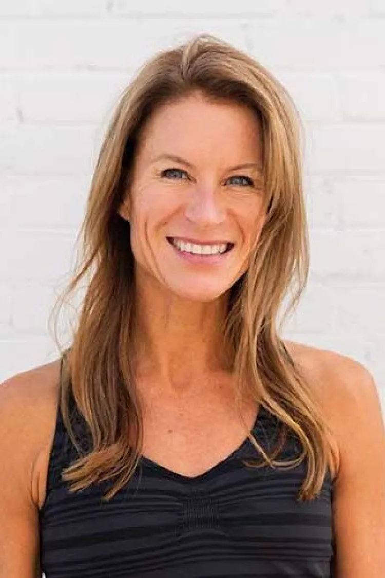 Smiling woman with long light brown hair in a black tank top standing in front of a white brick wall.
