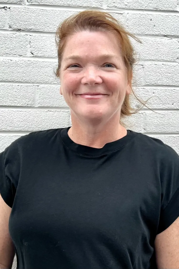 Woman with light brown hair in a black shirt smiling in front of a white brick wall.