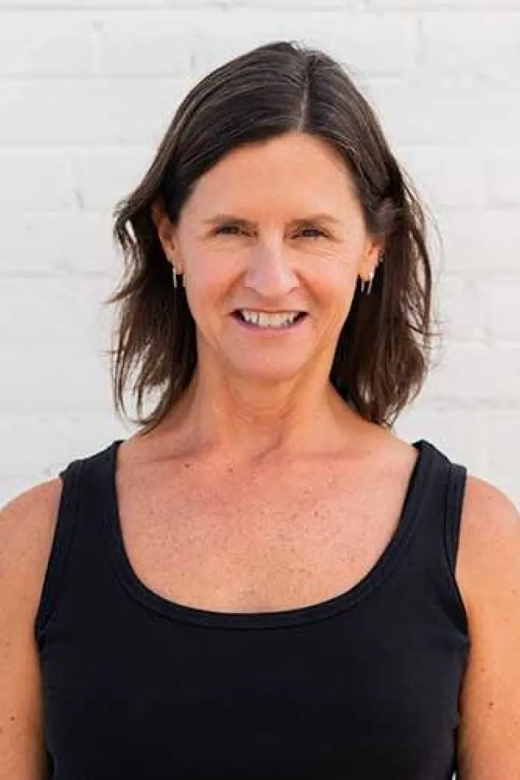 Woman with shoulder-length brown hair wearing a black tank top, smiling in front of a white brick wall.