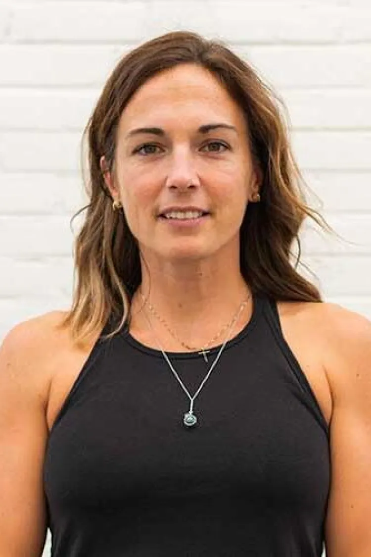 Woman with shoulder-length brown hair in a black tank top standing in front of a white brick wall.