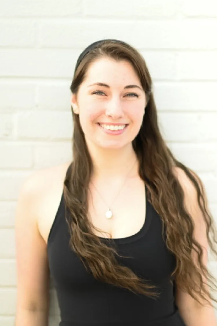 A young woman with long brown hair in a black tank top smiles in front of a light-colored brick wall.