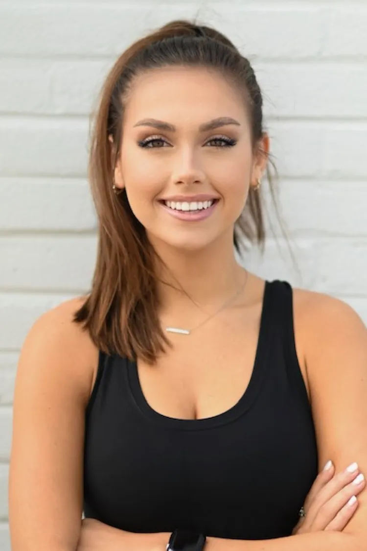 Woman with long brown hair in a ponytail, wearing a black tank top, smiling with arms crossed in front of a white wall.