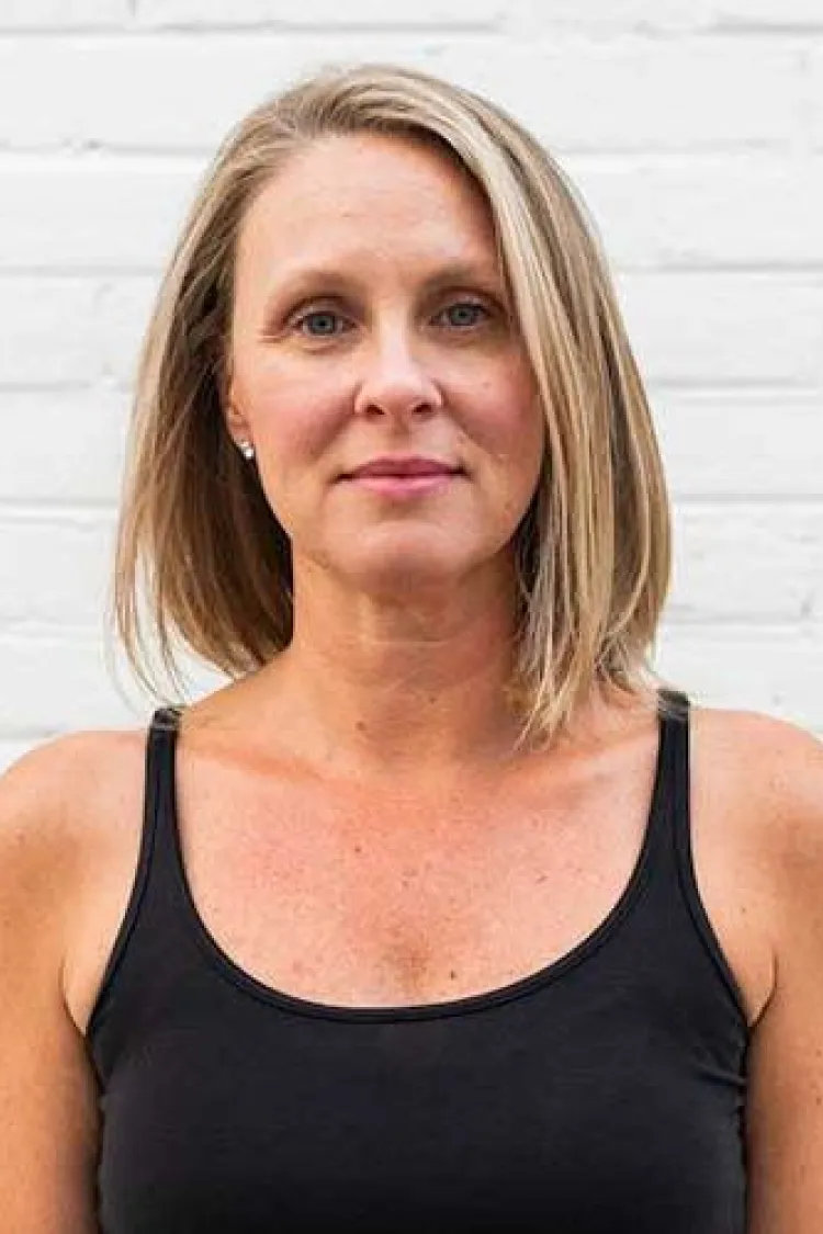 A woman with straight blonde hair wearing a black tank top stands in front of a white brick wall.