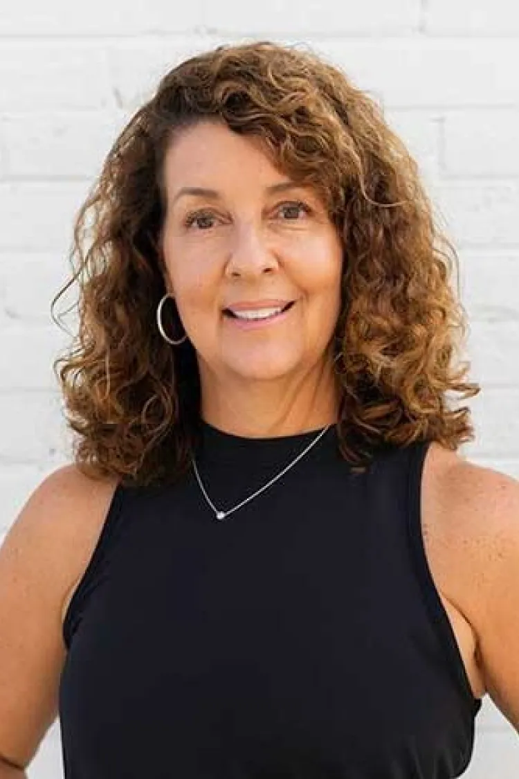 Smiling woman with curly brown hair wearing a sleeveless black top, hoop earrings, and a necklace.
