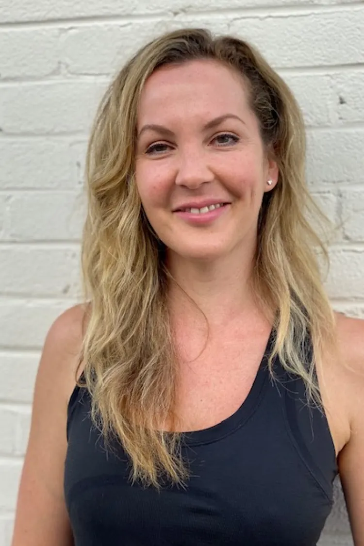 Smiling woman with wavy blonde hair in a black tank top standing in front of a white brick wall.
