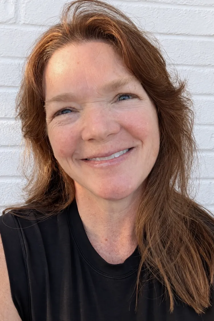 Smiling woman with long brown hair wearing a sleeveless black shirt stands in front of a white brick wall.